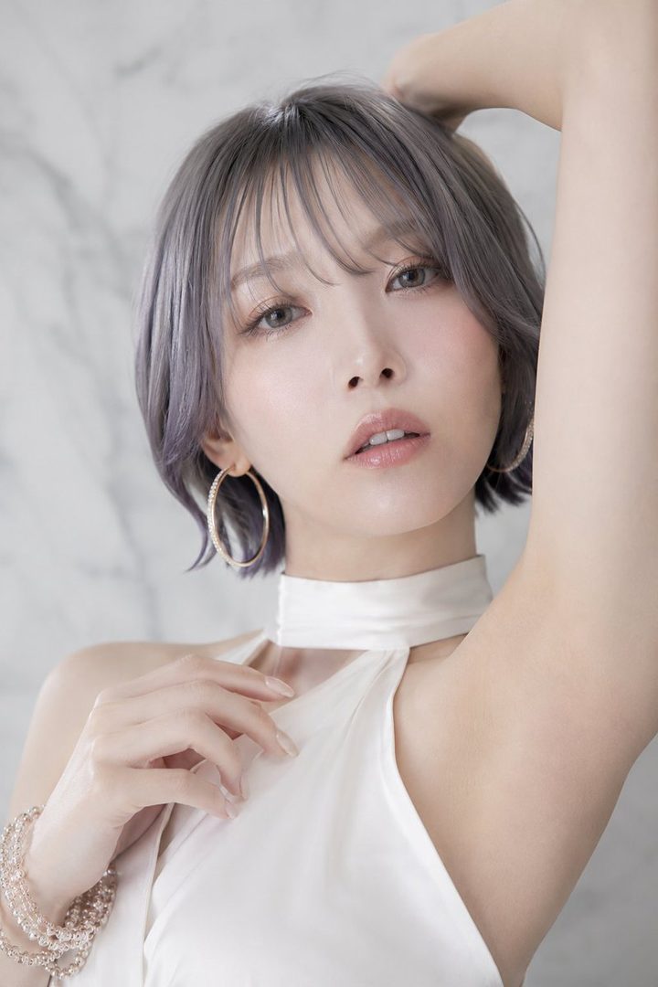 A woman with short silver hair and a white halter-neck dress poses elegantly indoors.