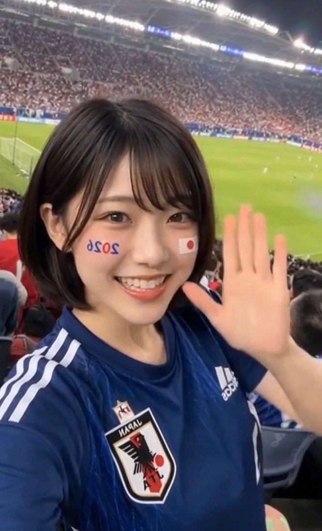 A young woman with short brown hair and bangs smiles at the camera, wearing a blue soccer jersey with face paint, at a stadium.