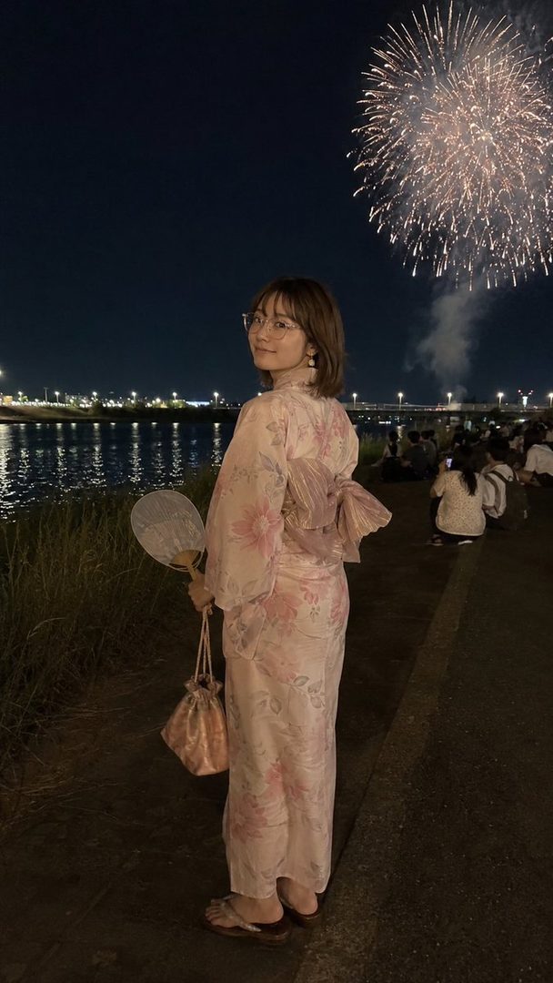 A young woman in a pink yukata with floral patterns stands by a river at night, looking over her shoulder at the camera, with fireworks exploding in the background.