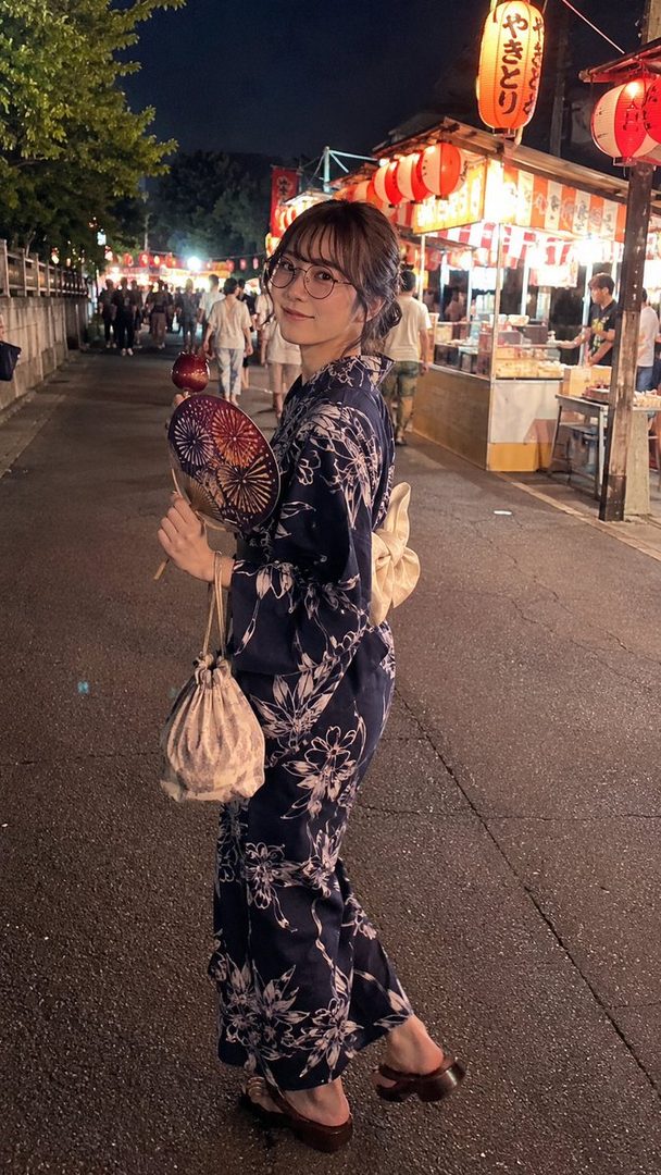 A young woman in a dark blue yukata with white floral patterns walks down a street at night, holding a fan and a small bag, with food stalls and lanterns in the background.