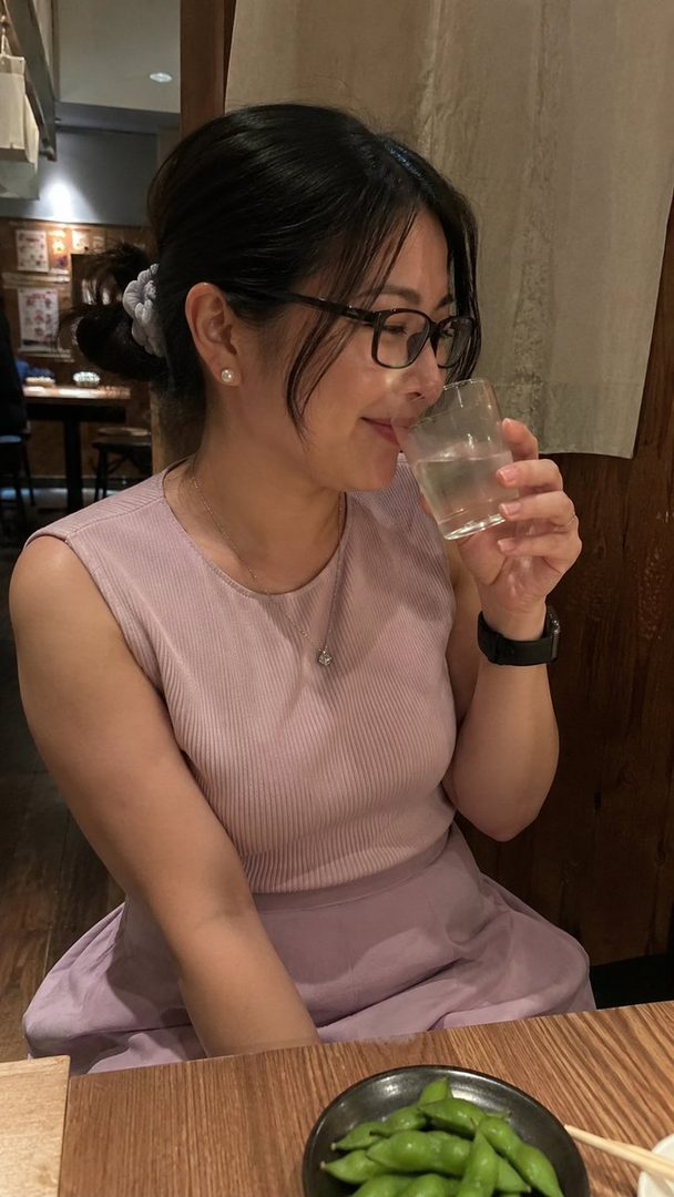A woman with black hair and glasses sips from a glass while sitting at a table with edamame.