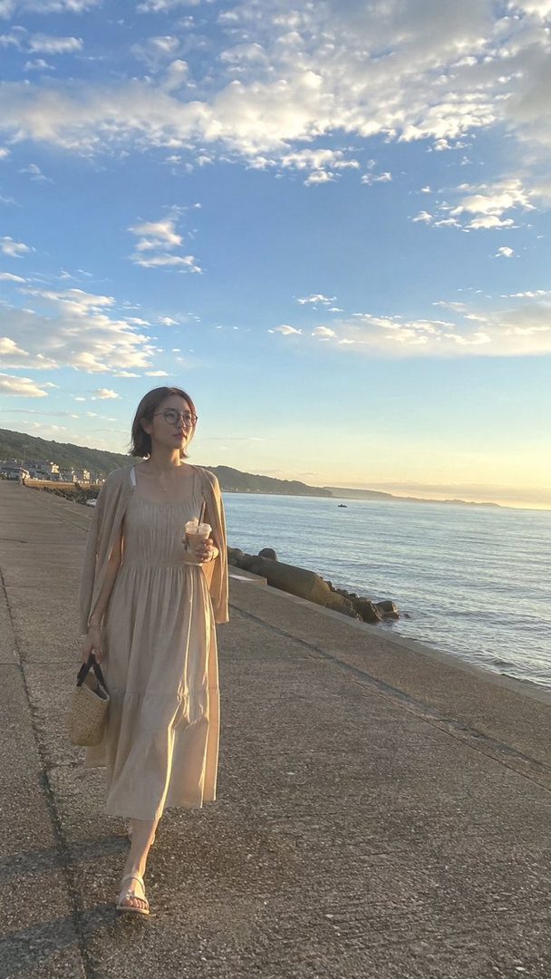 A woman in a light-colored dress walks along a concrete path by the sea at golden hour, holding an iced coffee.