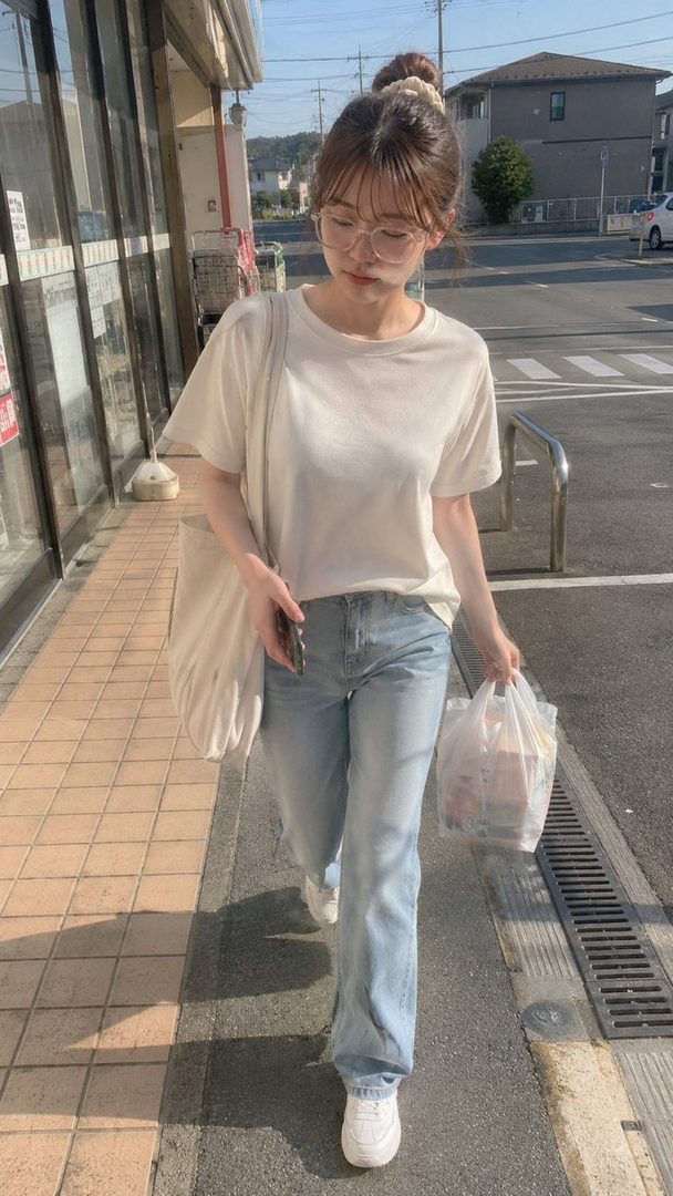 A young woman with glasses and a bun hairstyle walks down a sunny street, carrying a shopping bag.