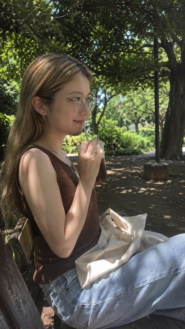 A young woman with long blonde hair and glasses sips a drink while sitting on a bench in a park on a sunny day.