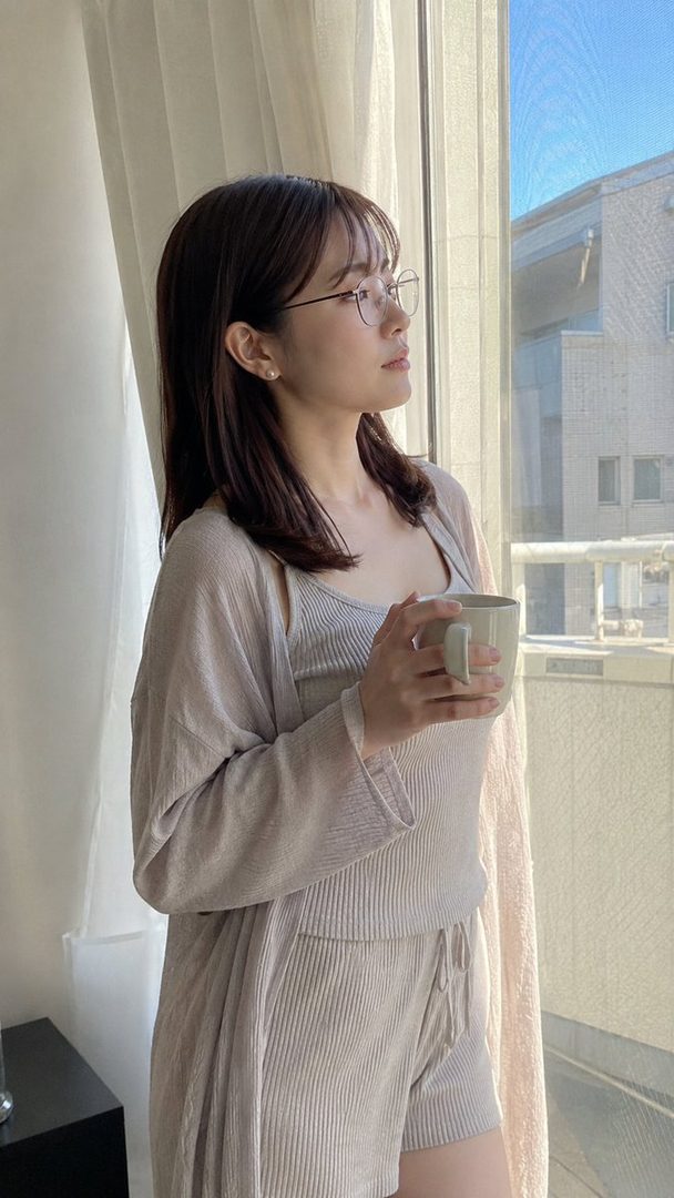A woman with brown hair and glasses, wearing a light knit top and shorts, stands by a window holding a mug and looking outside.