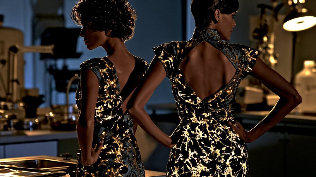 Two women in glowing, patterned dresses stand in an indoor setting, looking away from the camera.