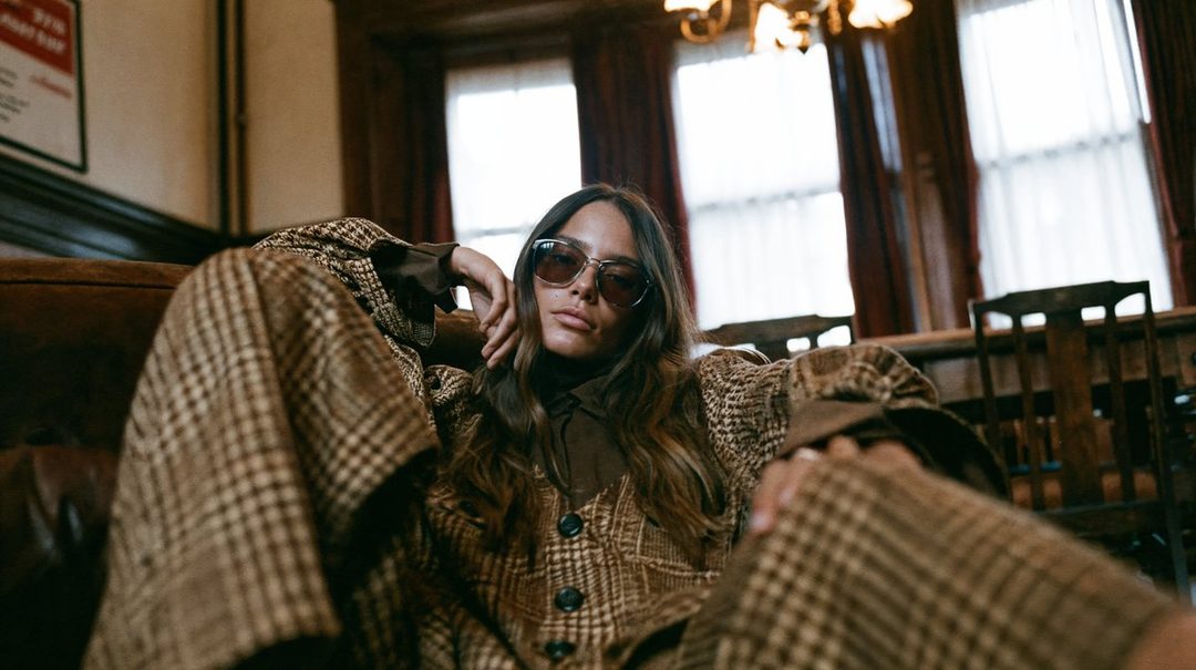 A woman with long brown hair and sunglasses wears a brown plaid suit while lounging indoors.