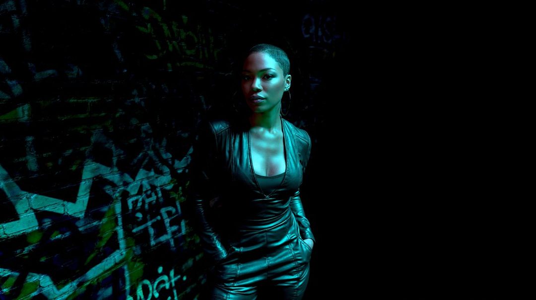 A woman with short black hair and a black leather outfit stands in a dark room with graffiti on the wall, illuminated by green and blue light.
