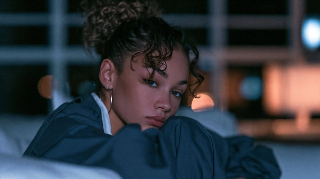 A photorealistic portrait of a young woman with curly brown hair tied up, wearing a dark blue casual top, looking directly at the camera with a serene expression in a dimly lit indoor setting.