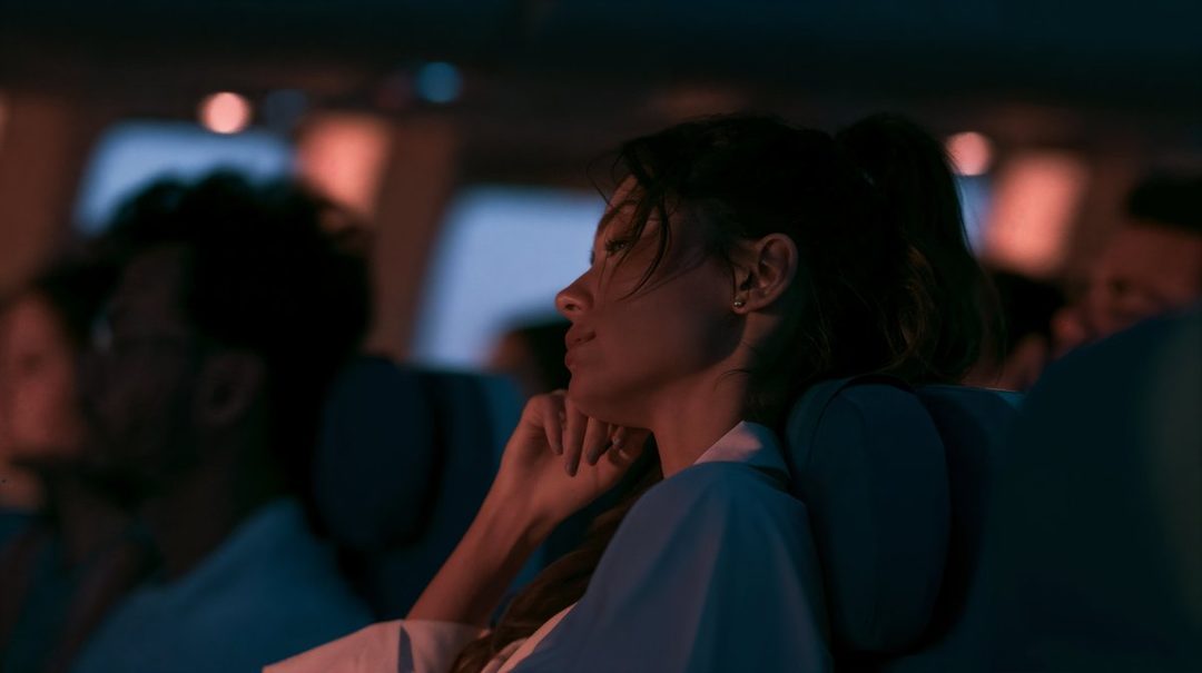 A woman with brown hair in a ponytail sits in a dimly lit cabin, looking out of a window with a serene expression.
