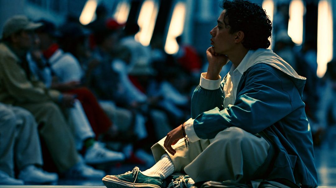 A young man with curly black hair, wearing a blue jacket and light pants, sits thoughtfully indoors with his chin resting on his hand.