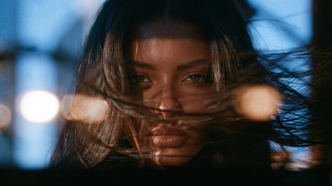A close-up photorealistic portrait of a woman with long brown hair, looking directly at the camera with a dramatic, cinematic lighting and bokeh in the background.
