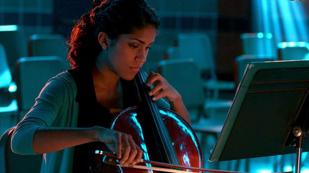 A young woman with curly dark hair plays the cello in a dimly lit room, illuminated by blue and orange light.