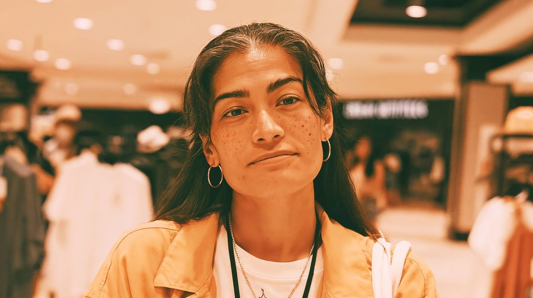 A photorealistic portrait of a young woman with long brown hair and freckles, wearing a casual yellow jacket and hoop earrings, looking directly at the camera in an indoor setting.