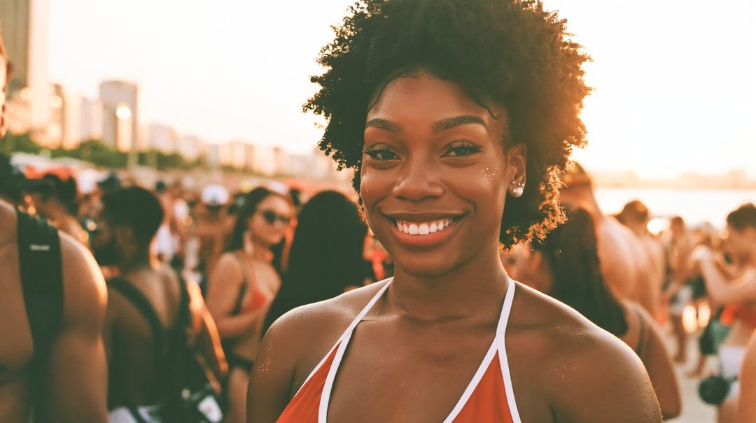 A smiling woman with short curly black hair wearing a red and white bikini top stands on a crowded beach during golden hour.