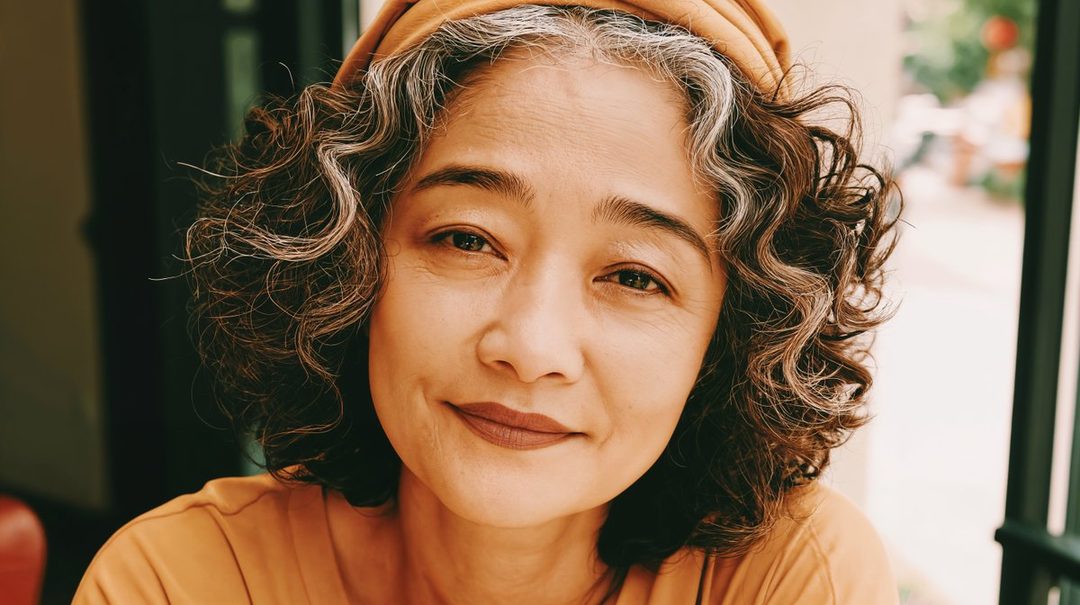 A close-up portrait of an East Asian woman with curly brown and silver hair, wearing a casual top and a headscarf, looking serenely at the camera.