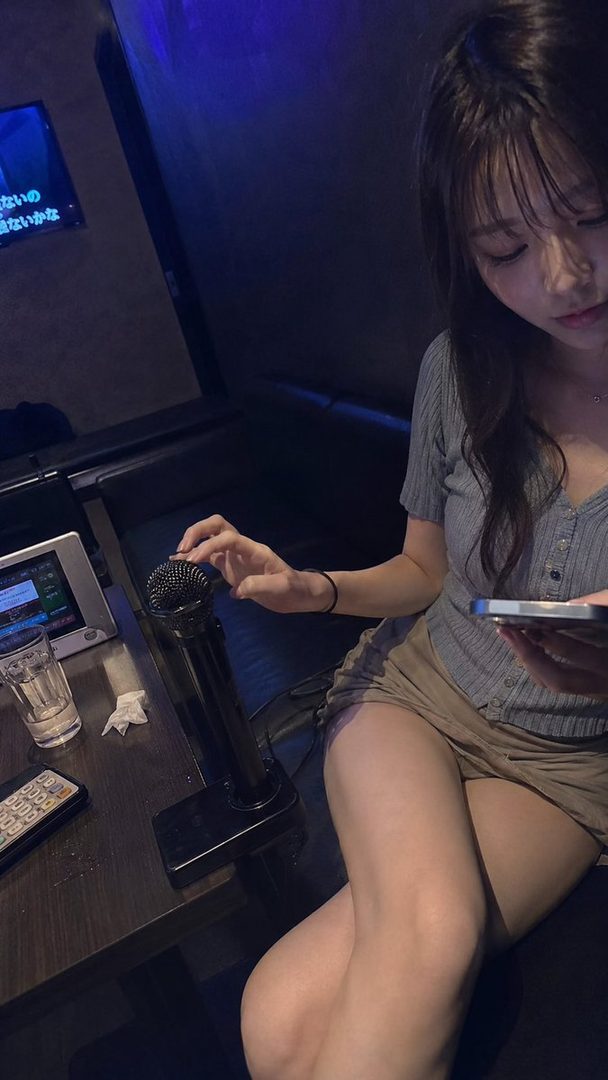 A young woman with long brown hair and bangs, wearing a grey top and shorts, sits indoors holding a microphone and a smartphone.