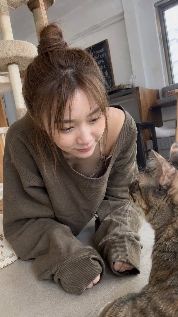 A young woman with brown hair tied in a bun and bangs, wearing a loose olive green sweater, is on the floor interacting with a cat in an indoor setting.