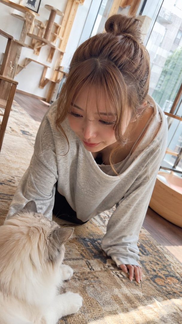 A young woman with brown hair in a bun and bangs, wearing a grey long-sleeved top, looks down at a cat on a patterned rug indoors.