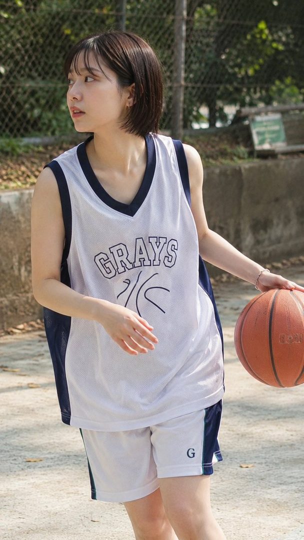 A young woman with short brown hair wears a white and blue basketball jersey and shorts, holding a basketball outdoors.