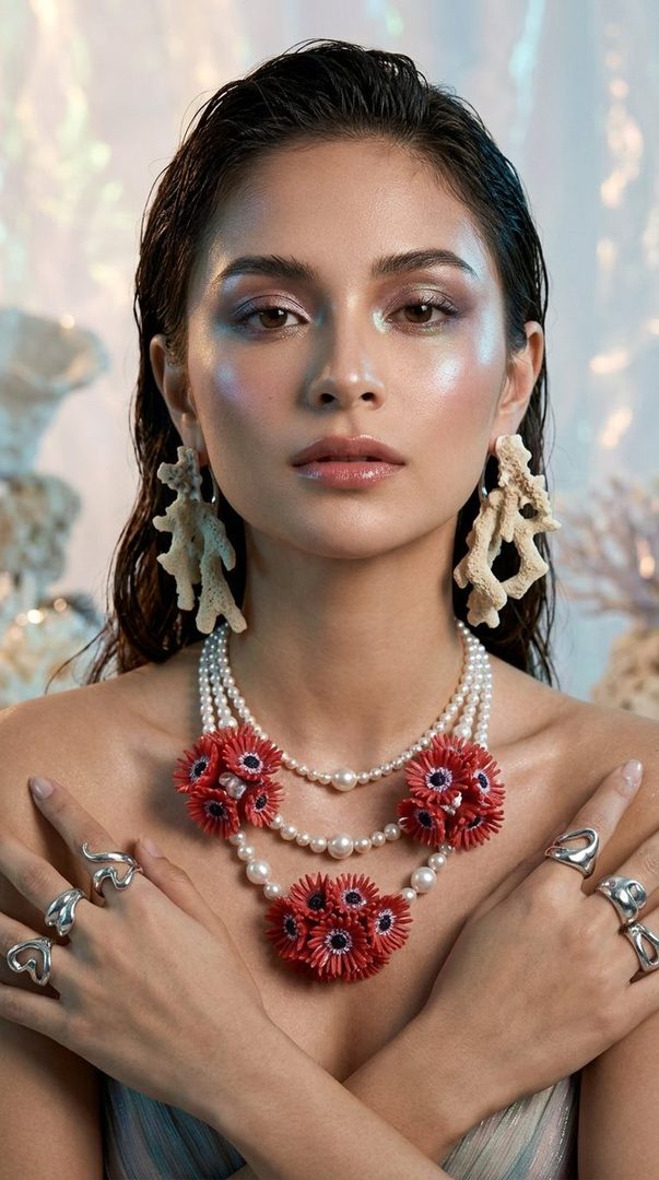 A woman with wet brown hair and coral-inspired jewelry, including a pearl and red floral necklace, poses for a fashion shot.