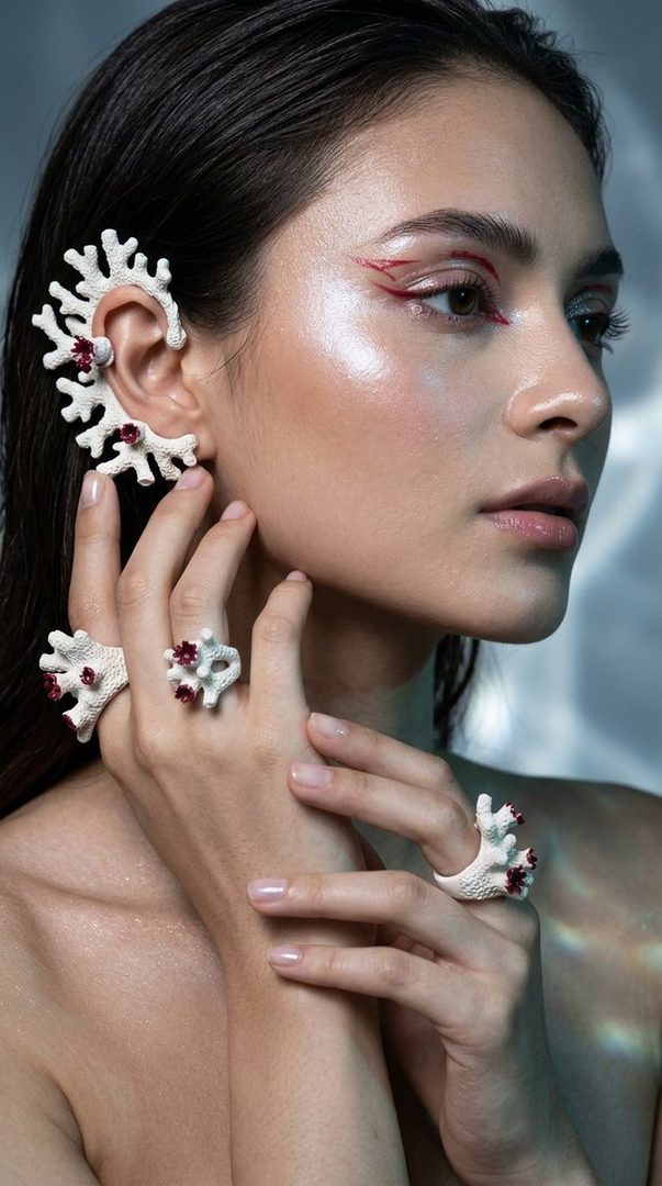 A woman with dark hair and red eye makeup wears white coral-inspired jewelry, including an ear cuff and rings, in a fashion magazine style shot.
