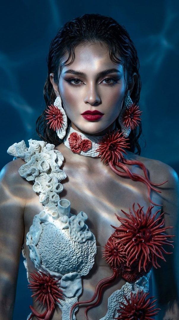 A woman with dark hair and red lipstick wears an elaborate coral-inspired outfit and jewelry against a blue background with water reflections.