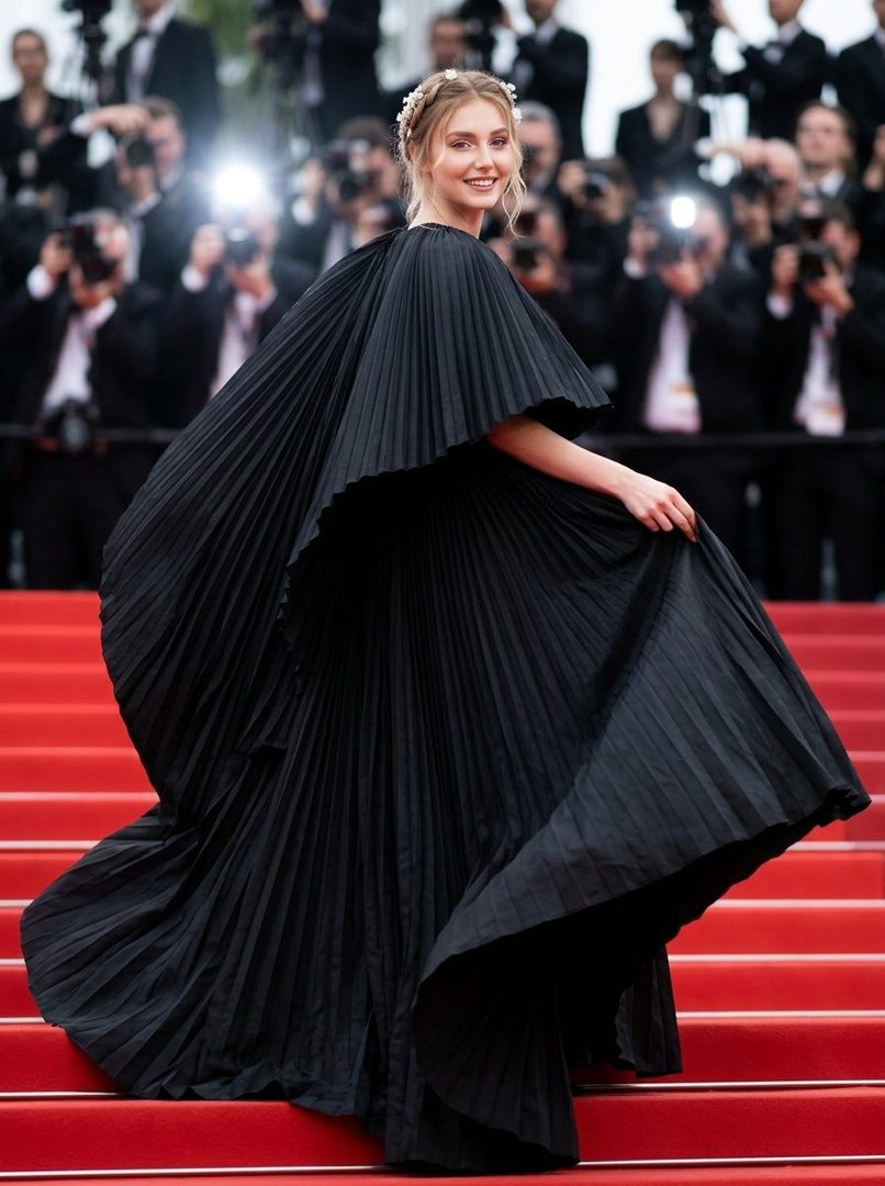 A woman in a flowing black pleated gown walks on a red carpet, smiling at the camera with photographers in the background.