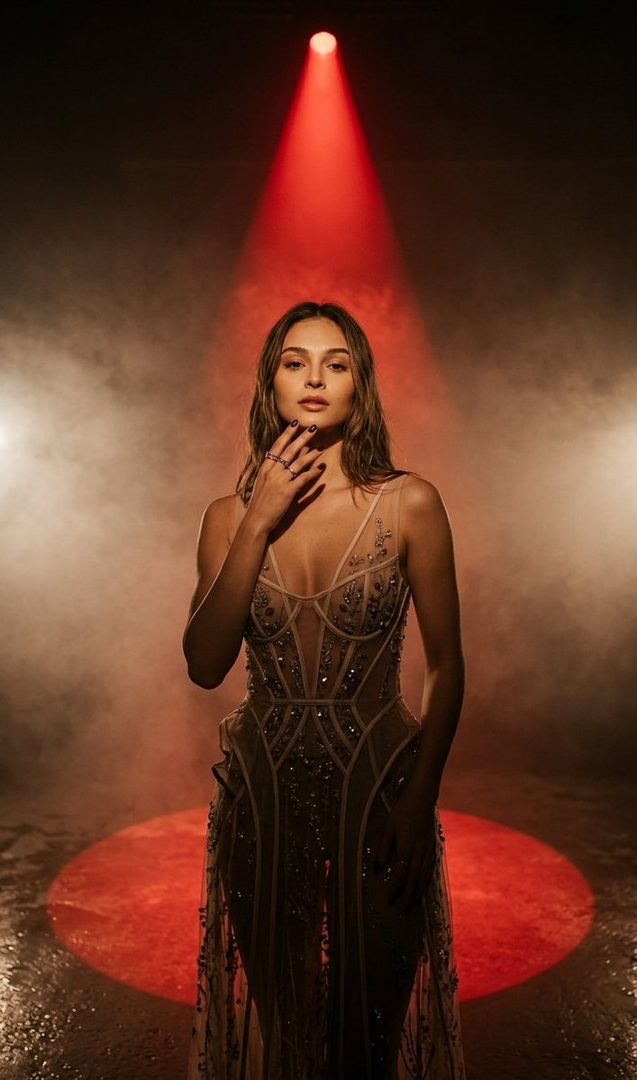 A woman in a sheer, embellished dress stands under a red spotlight in a smoky studio.