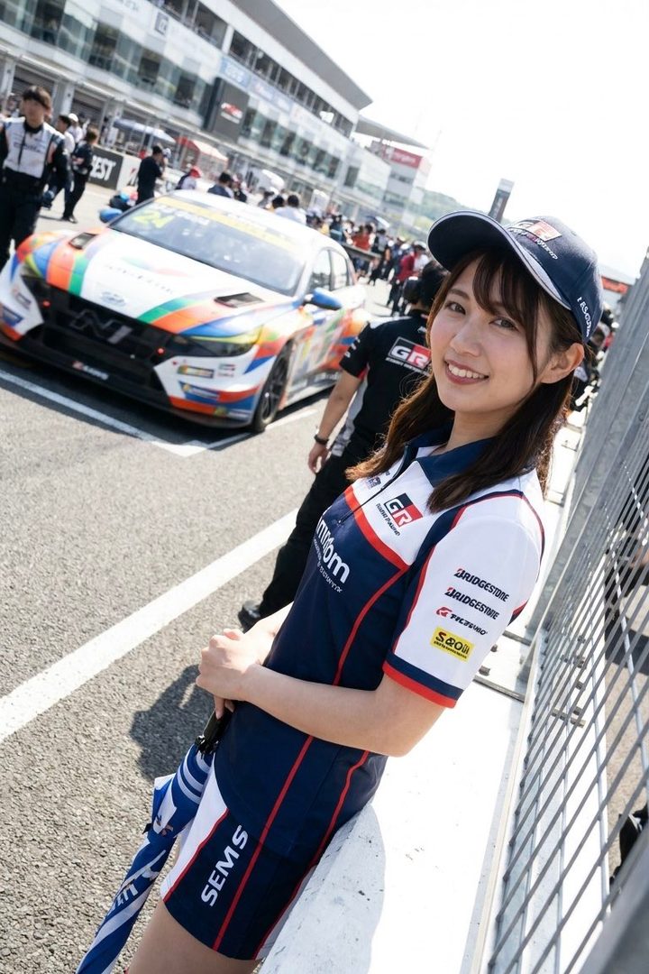 A smiling woman in a race queen uniform and cap stands next to a race car on a track.