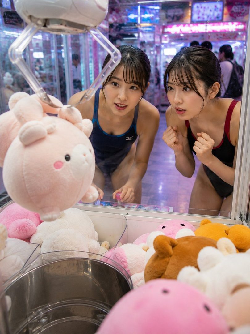 Two young women in swimsuits are intently watching a claw machine in an arcade, with one of them operating the joystick.