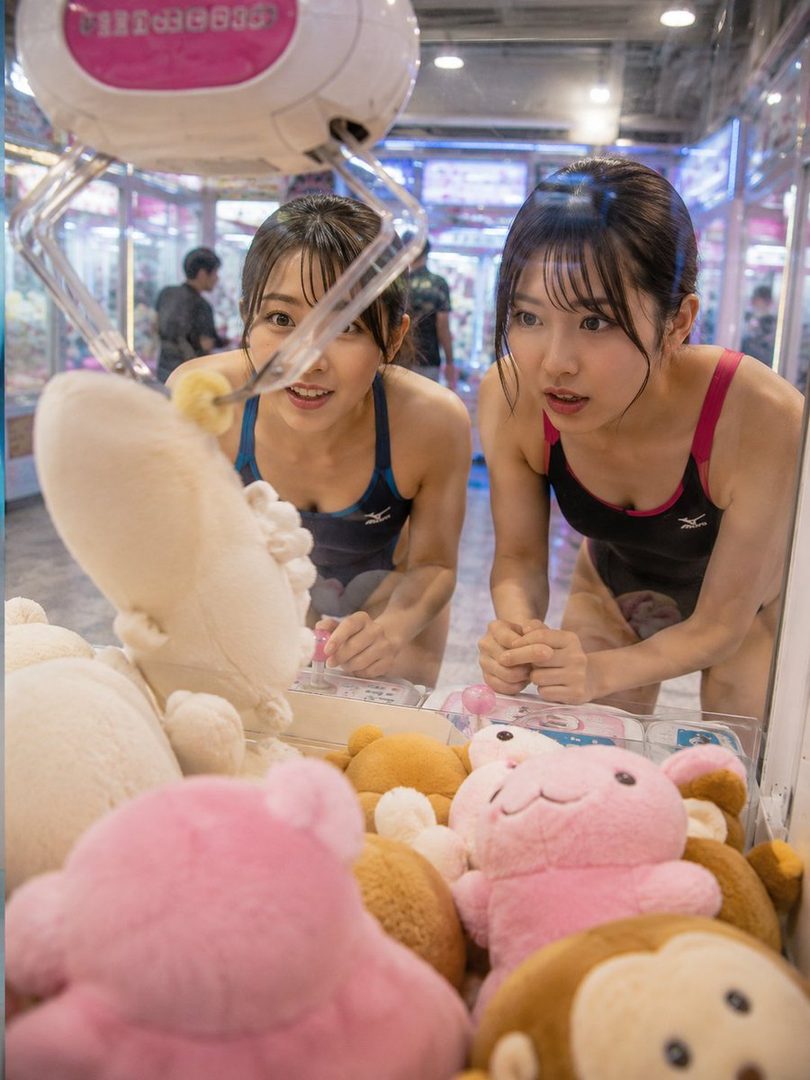 Two young women in swimsuits are intently playing a claw machine filled with plush toys.