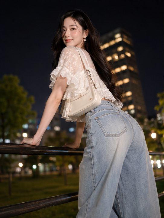 A young woman with long dark hair and a white lace crop top leans on a railing at night in an urban park, looking back at the camera with a gentle smile.