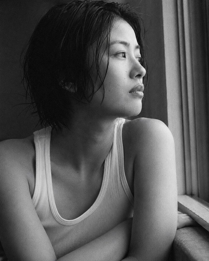 A black and white photorealistic portrait of a Japanese woman with short, wet-looking black hair, wearing a white ribbed tank top, looking out a window with a neutral expression.