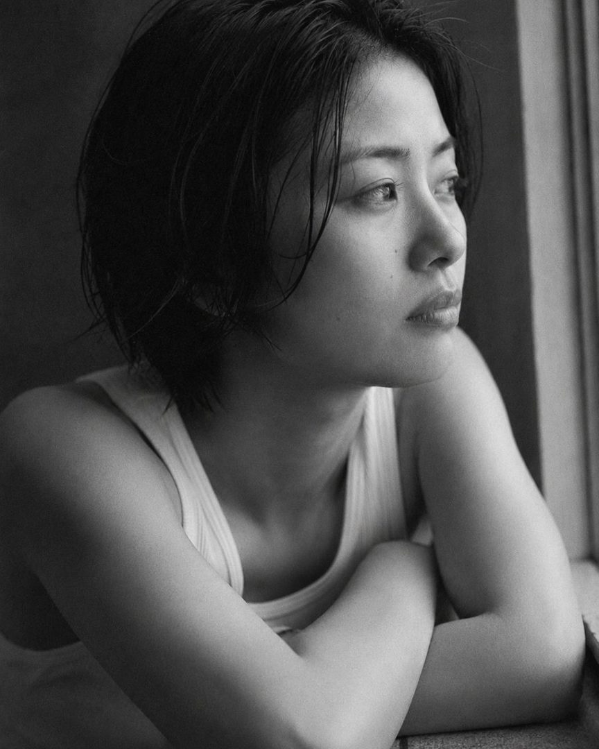 A black and white photorealistic portrait of a Japanese woman with short, wet-looking black hair, wearing a tank top, looking out a window with a serene expression.
