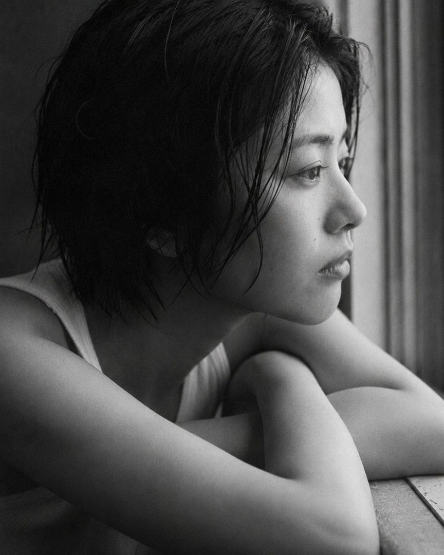 A black and white photorealistic portrait of a Japanese woman with short, wet-looking black hair, looking out a window with a serene expression.