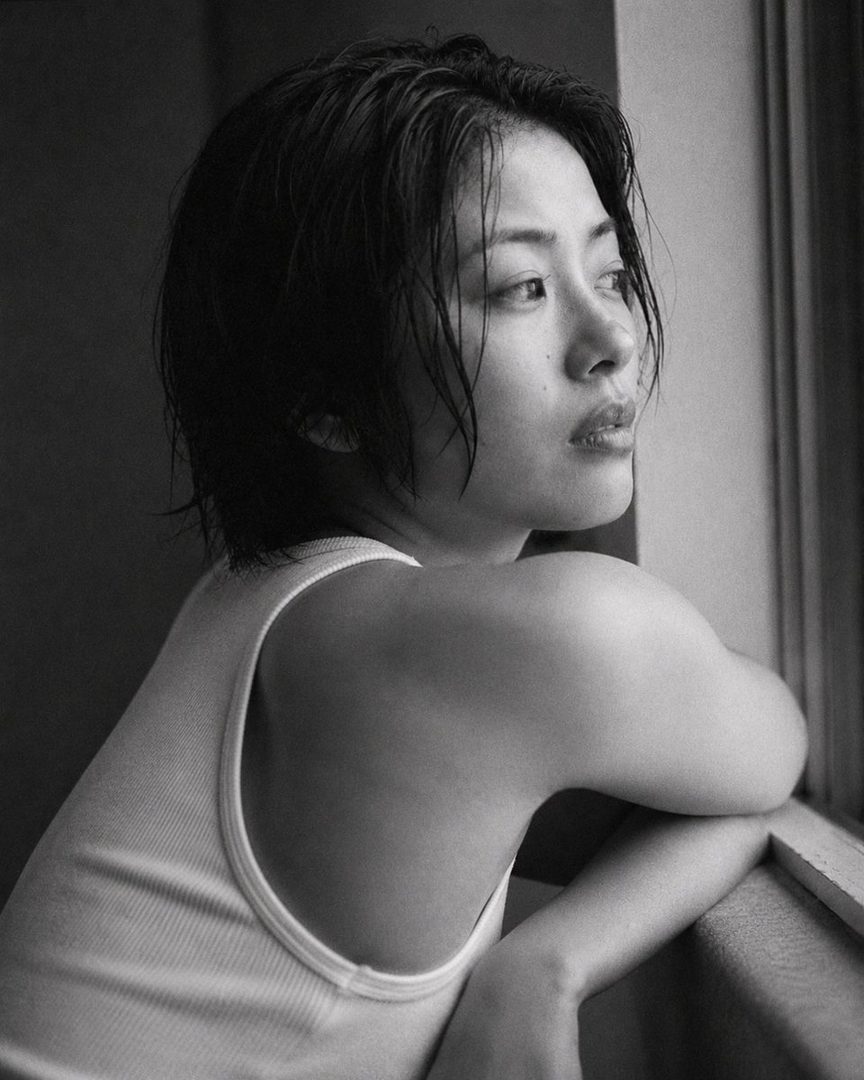 A black and white photorealistic portrait of a Japanese woman with short, wet-looking black hair, wearing a white tank top, looking out a window.