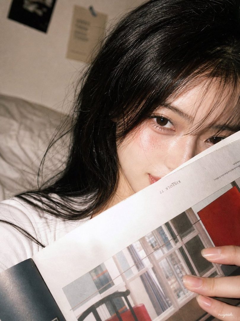 A close-up portrait of a young woman with long black hair, partially obscured by a magazine she is holding, looking directly at the camera.