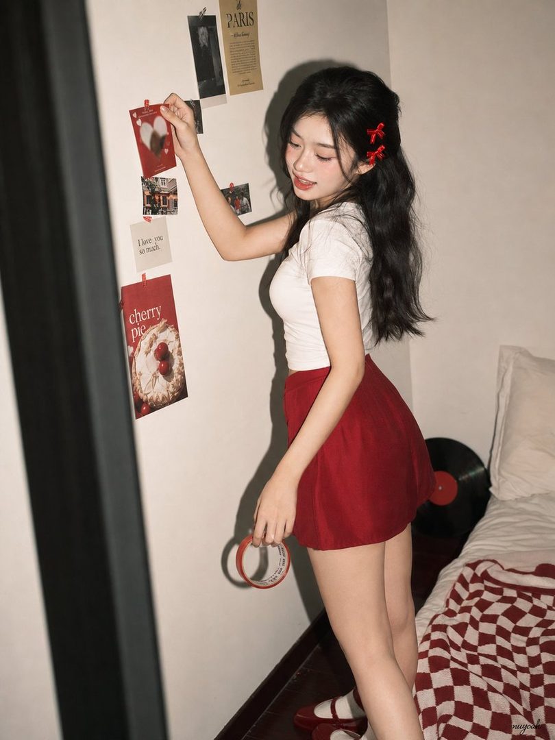 A young woman in a white top and red skirt is decorating a wall with pictures in a cozy indoor setting.