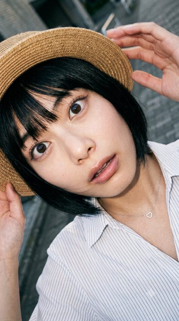 A close-up portrait of a young woman with short black hair and bangs, wearing a straw hat and a striped blouse, looking directly at the camera with wide eyes.