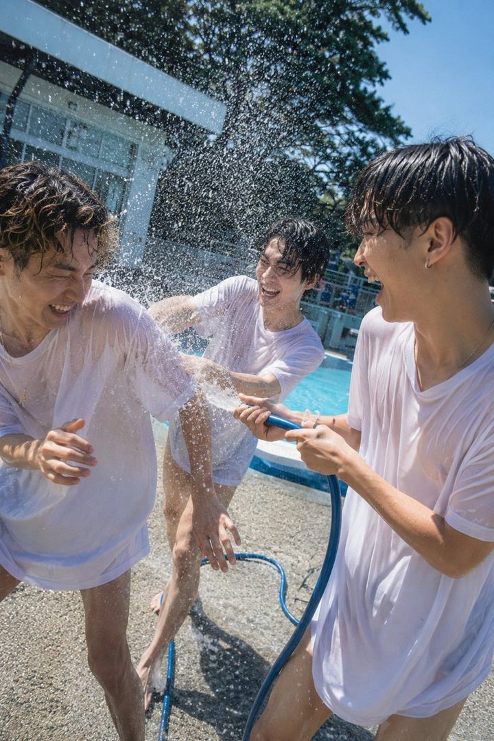 Three young men in white t-shirts are playfully splashing water on each other with a hose by a swimming pool on a sunny day.