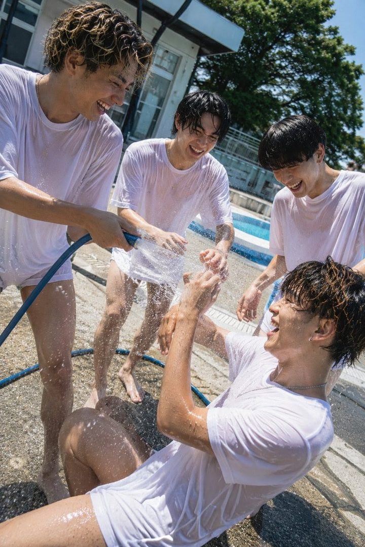 Four young men are happily playing with water from a hose outdoors on a sunny day, all wearing wet white t-shirts.