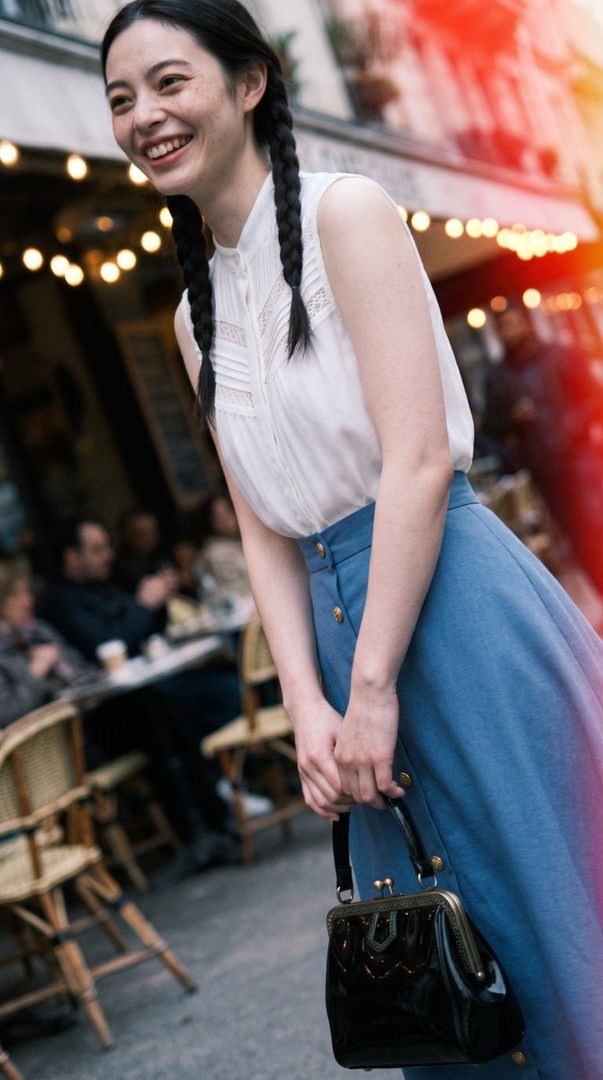 A smiling woman with black braided hair and freckles, wearing a white blouse and blue skirt, stands on a city street with a cafe in the background.