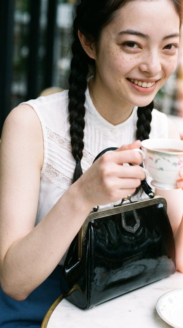 A smiling young woman with black braided hair and freckles sits at a cafe table, holding a teacup and a black handbag.