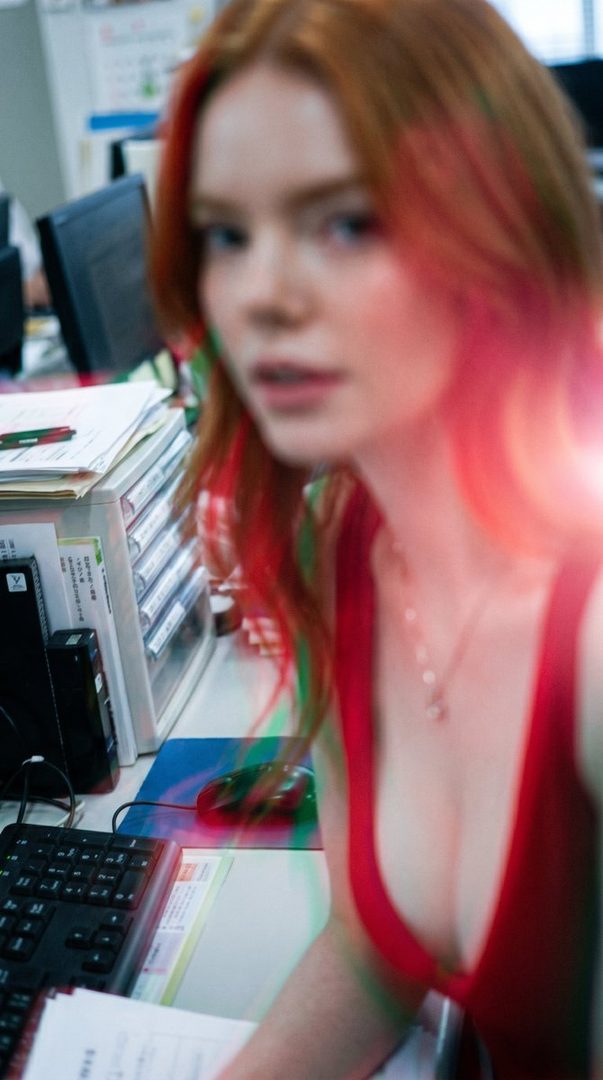 A photorealistic portrait of a woman with long red hair wearing a red top, sitting at an office desk with a keyboard and mouse.