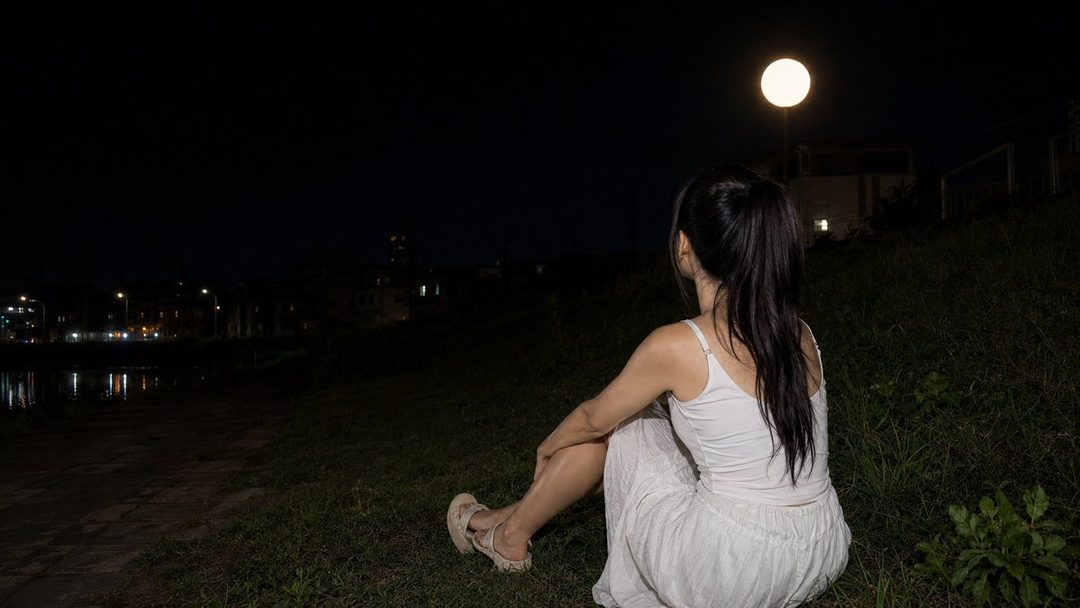 A woman with long black hair in a ponytail sits on a grassy bank at night, looking towards a bright street light that resembles a full moon.