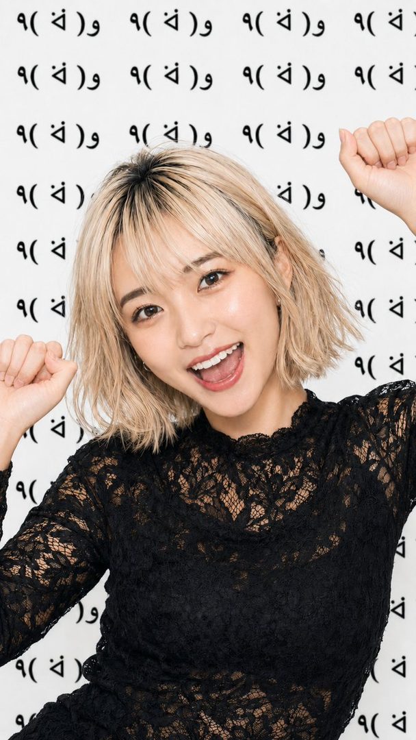 A cheerful woman with blonde short hair and bangs, wearing a black lace top, smiles brightly at the camera with her fists raised against a white background with repeating black symbols.