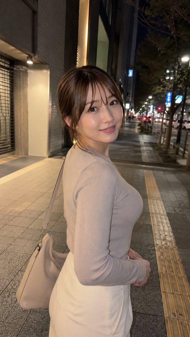 A young woman with brown hair and bangs smiles at the camera while walking on a city street at night.