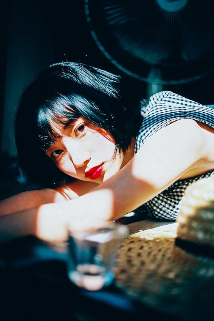 A young woman with short black hair and red lipstick lies on the floor, bathed in sunlight, with a fan in the background.