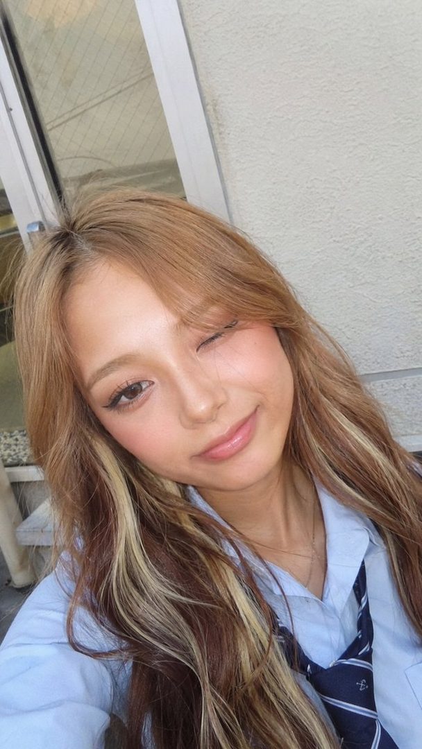 A young woman with blonde and brown streaked hair, wearing a light blue shirt and a dark blue tie, winks at the camera in a selfie.